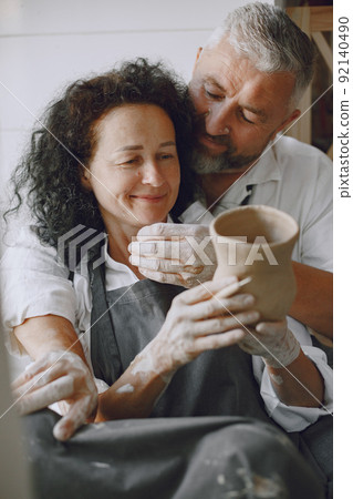 Mutual creative work. Adult elegant couple in casual clothes and aprons. People creating a bowl on a pottery wheel in a clay studio. Mutual creative work. Adult elegant couple in casual clothes and aprons. People creating a bowl on a pottery wheel in a clay studio. 92140490