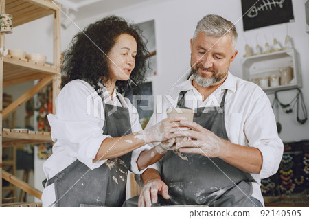 Mutual creative work. Adult elegant couple in casual clothes and aprons. People creating a bowl on a pottery wheel in a clay studio. 92140505
