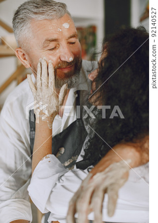 Mutual creative work. Adult elegant couple in casual clothes and aprons. People creating a bowl on a pottery wheel in a clay studio. Mutual creative work. Adult elegant couple in casual clothes and aprons. People creating a bowl on a pottery wheel in a clay studio. 92140527