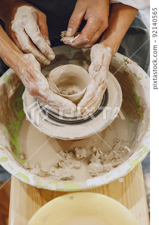 Mutual creative work. Adult elegant couple in casual clothes and aprons. People creating a bowl on a pottery wheel in a clay studio. Mutual creative work. Adult elegant couple in casual clothes and aprons. People creating a bowl on a pottery wheel in a clay studio. 92140565