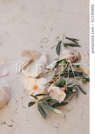 Summer wedding floral composition. Closeup of apricot oleander blossom, flowers and green olive tree branches. Beige marble background with silk ribbon. Blurred background, selective focus. Vertical. 92140866