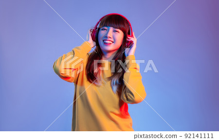 Young asian woman in yellow sweatshirt wearing red headphones listen to music on the purple screen background.. 92141111