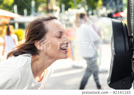 Profile side close-up view young adult beautiful woman enjoy fresh moisturized air blowing from big cooling fan machine hot sunny summer weather outdoors. Mist spraying ventilator system city street 92141185