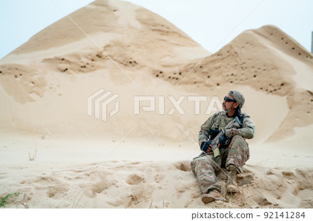 Military or soldier man with sunglasses sit on sand and look around in area look like desert Military or soldier man with sunglasses sit on sand and look around in area look like desert 92141284