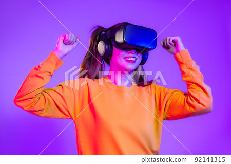 Metaverse concept. Young asian woman in orange sweatshirt wearing vr headset posing touching playing on the purple screen background.. Metaverse concept. Young asian woman in orange sweatshirt wearing vr headset posing touching playing on the purple screen background.. 92141315