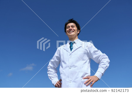 A man in a white coat looking forward with a smile with his hands on his hips under the blue sky A man in a white coat looking forward with a smile with his hands on his hips under the blue sky 92142173