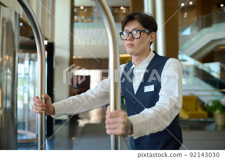 Young man in uniform of bellboy pushing cart with luggage along lounge 92143030