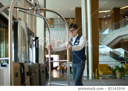 Young bellboy in uniform pushing cart with suitcases of hotel clients or guests Young bellboy in uniform pushing cart with suitcases of hotel clients or guests 92143034