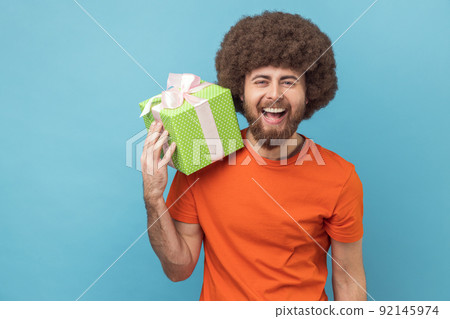 Portrait of extremely happy man with Afro hairstyle wearing orange T-shirt holding green present box and looking at camera with joyful expression. Indoor studio shot isolated on blue background. 92145974
