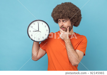 Portrait of confused puzzled man with Afro hairstyle wearing orange T-shirt holding big wall clock and holding chin, thinks about deadline. Indoor studio shot isolated on blue background. 92145981