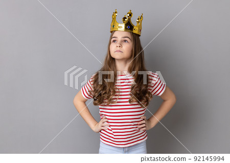 Portrait of confident little girl princess wearing striped T-shirt keeping hands on hips, self-motivation and dreams to be best. Indoor studio shot isolated on gray background. 92145994