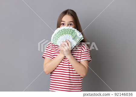 Portrait of charming little girl wearing striped T-shirt hiding half of face behind fan of hundred euro bills, enjoying smell of money. Indoor studio shot isolated on gray background. 92146036
