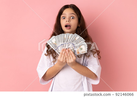 Portrait of shocked rich little girl wearing white T-shirt holding big fan of dollar banknotes, looking at camera with surprised expression. Indoor studio shot isolated on pink background. 92146066