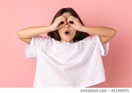 Little girl wearing white T-shirt making glasses shape with fingers, looking through binoculars hand gesture and expressing shock, astonishment. Indoor studio shot isolated on pink background. 92146093