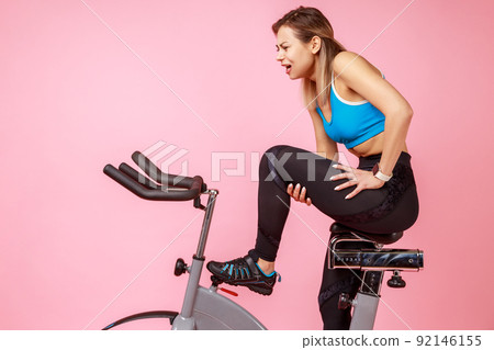 Side view portrait of athletic woman has workout at home, riding exercise bike and injured her leg, feeling pain in thigh, wearing sports tights and top. Indoor studio shot isolated on pink background Side view portrait of athletic woman has workout at home, riding exercise bike and injured her leg, feeling pain in thigh, wearing sports tights and top. Indoor studio shot isolated on pink background 92146155