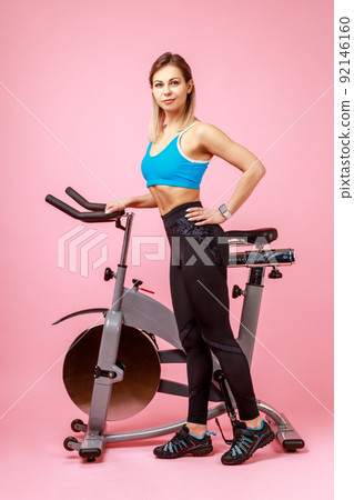 Full length portrait of charming woman with slim body shape posing near exercise bike and looking at camera, wearing sports tights and top. Indoor studio shot isolated on pink background. Full length portrait of charming woman with slim body shape posing near exercise bike and looking at camera, wearing sports tights and top. Indoor studio shot isolated on pink background. 92146160
