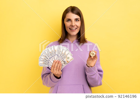 Portrait of optimistic dark haired woman holding bitcoin and big fan of dollar bills, exchange crypto currency, wearing purple hoodie. Indoor studio shot isolated on yellow background. 92146196