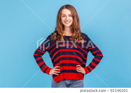 Portrait of smiling positive woman wearing striped casual style sweater looking at camera, keeping hands on hips, having confident expression. Indoor studio shot isolated on blue background. Portrait of smiling positive woman wearing striped casual style sweater looking at camera, keeping hands on hips, having confident expression. Indoor studio shot isolated on blue background. 92146201