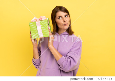 Portrait of curious dark haired woman holding shaking green wrapped present box, thinks what is inside, wearing purple hoodie. Indoor studio shot isolated on yellow background. 92146243