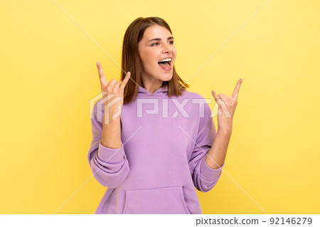 Excited enthusiastic woman with dark hair showing rock and roll gesture. screaming rapturously, looking away, wearing purple hoodie. Indoor studio shot isolated on yellow background. 92146279