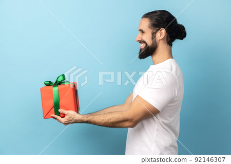 Side view of handsome delighted man with beard wearing white T-shirt giving wrapped gift box and smiling, looking ahead, happy holiday. Indoor studio shot isolated on blue background. 92146307