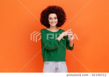 Internet trends. Smiling woman with Afro hairstyle wearing green sweater crossing fingers to make hashtag sign and looking at camera with happiness. Indoor studio shot isolated on orange background. 92146312