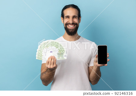 Portrait of man with beard wearing white T-shirt holding fan of euro banknotes and smart phone with black blank screen, making money online. Indoor studio shot isolated on blue background. 92146316