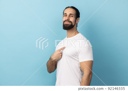 Portrait of self confident man with beard wearing white T-shirt pointing at himself, feeling proud and self-important, having big ego. Indoor studio shot isolated on blue background. 92146335
