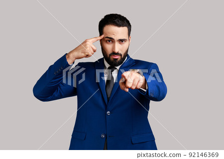 Displeased bearded man holding finger near temple and pointing on you, showing stupid gesture, mocking, wearing official style suit. Indoor studio shot isolated on gray background. 92146369
