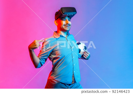 Happy smiling man in shirt wearing virtual reality glasses, standing with satisfied expression, pointing at himself with proud. Indoor studio shot isolated on colorful neon light background. 92146423
