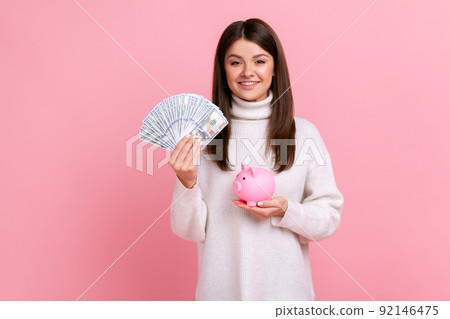 Optimistic brunette female holding fan of dollar bills and piggy bank, savings, growth of deposit, wearing white casual style sweater. Indoor studio shot isolated on pink background. 92146475