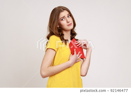 Love letter on Valentine's day. Portrait of teen girl in yellow casual T- shirt hugging letter in red envelope, holding greeting card with expression. Indoor studio shot isolated on gray background. 92146477