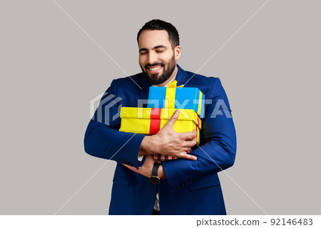 Satisfied bearded man with toothy smile embracing stack of present boxes, keeping eyes closed, enjoying holiday, wearing official style suit. Indoor studio shot isolated on gray background. 92146483