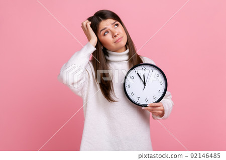 Pensive confused female with dark hair holding big wall clock, looking away, keeping hand on head, wearing white casual style sweater. Indoor studio shot isolated on pink background. Pensive confused female with dark hair holding big wall clock, looking away, keeping hand on head, wearing white casual style sweater. Indoor studio shot isolated on pink background. 92146485