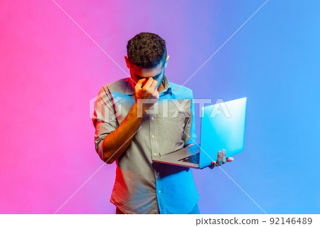 Portrait of tired man in shirt holding portable computer in hands and rubbing his eyes, being exhausted working long hours. Indoor studio shot isolated on colorful neon light background. Portrait of tired man in shirt holding portable computer in hands and rubbing his eyes, being exhausted working long hours. Indoor studio shot isolated on colorful neon light background. 92146489