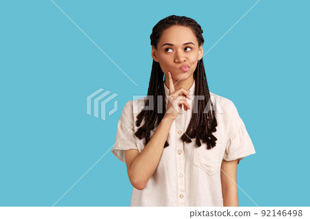 Portrait of thoughtful woman with black dreadlocks keeps index finger on cheek, considers something, has pensive expression, wearing white shirt. Indoor studio shot isolated on blue background. 92146498