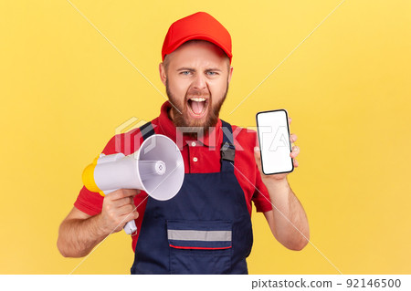 Portrait of angry bearded worker man holding megaphone and blank phone display with advertisement area, screaming with anger, wearing blue uniform. Indoor studio shot isolated on yellow background. 92146500