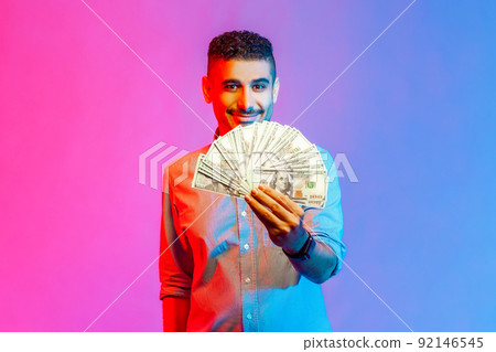 Portrait of rich handsome man in shirt holding dollars, looking at camera with toothy smile, being happy to win lottery. Indoor studio shot isolated on colorful neon light background. Portrait of rich handsome man in shirt holding dollars, looking at camera with toothy smile, being happy to win lottery. Indoor studio shot isolated on colorful neon light background. 92146545
