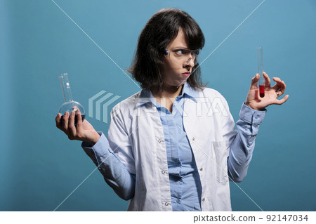 Upset angry professional laboratory chemist holding chemistry glass jars filled with chemical unidentified compounds. Biochemistry specialist having glass flasks filled with liquid substances. Upset angry professional laboratory chemist holding chemistry glass jars filled with chemical unidentified compounds. Biochemistry specialist having glass flasks filled with liquid substances. 92147034