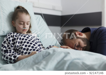 Worried father sitting next to ill little girl resting on patient bed inside hospital pediatrics ward room. Unwell sick kid sleeping while uneasy parent sitting beside her inside healthcare facility. Worried father sitting next to ill little girl resting on patient bed inside hospital pediatrics ward room. Unwell sick kid sleeping while uneasy parent sitting beside her inside healthcare facility. 92147146