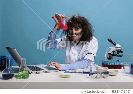 Foolish funny looking crazy chemist having glass flask filled with experimental liquid serum after failed chemical experiment. Mad silly lab worker having wild look and weird face expression. 92147147
