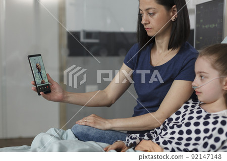 Ill kid and parent discussing with physician on smartphone virtual online call. Mother and sick daughter talking with doctor on telehealth phone videocall while sitting in hospital pediatrics ward. Ill kid and parent discussing with physician on smartphone virtual online call. Mother and sick daughter talking with doctor on telehealth phone videocall while sitting in hospital pediatrics ward. 92147148