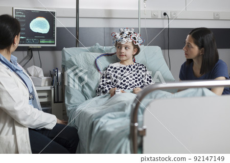 Hospitalized sick girl wearing EEG headset resting in patient bed while doctor analyzing brain condition. Neurology specialist examining neurological disease state of ill kid. Hospitalized sick girl wearing EEG headset resting in patient bed while doctor analyzing brain condition. Neurology specialist examining neurological disease state of ill kid. 92147149