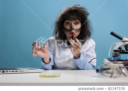Dumb crazy looking mad chemist smelling experimental liquid red serum while sitting at desk in laboratory. Insane scientist with wacky hair and dirty face holding tube filled with chemical compounds. Dumb crazy looking mad chemist smelling experimental liquid red serum while sitting at desk in laboratory. Insane scientist with wacky hair and dirty face holding tube filled with chemical compounds. 92147150
