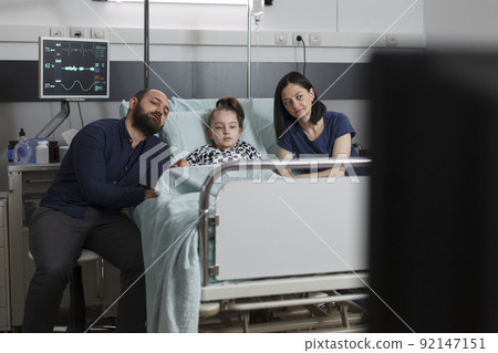 Caring parents sitting beside hospitalized daughter while watching television show inside children healthcare facility. Mother and father resting with sick little girl while enjoying family tv series. Caring parents sitting beside hospitalized daughter while watching television show inside children healthcare facility. Mother and father resting with sick little girl while enjoying family tv series. 92147151