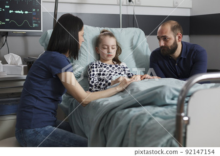 Worried parents sitting beside sick daughter hospitalized in pediatric healthcare facility. Sleeping ill little girl comforted by caring mother and father while resting in patient bed. Worried parents sitting beside sick daughter hospitalized in pediatric healthcare facility. Sleeping ill little girl comforted by caring mother and father while resting in patient bed. 92147154