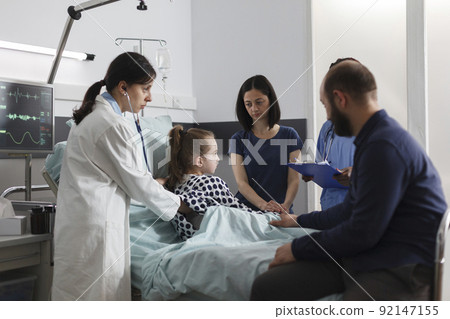 Expert pediatrist checking sick girl health condition using stethoscope while parents sitting besider her. General practitioner consulting ill little girl under treatment inside clinic pediatric ward. 92147155