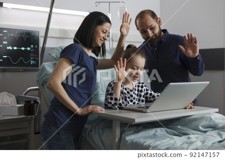 Ill little girl under treatment talking with relatives on online call while sitting with loving parents inside hospital ward. Mother and father standing beside daughter while waving at videocall. Ill little girl under treatment talking with relatives on online call while sitting with loving parents inside hospital ward. Mother and father standing beside daughter while waving at videocall. 92147157