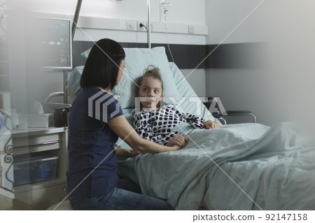 Mother sitting beside hospitalized sick daughter resting in hospital pediatric ward. Caring woman comforting ill little girl under treatment inside children healthcare facility room. Mother sitting beside hospitalized sick daughter resting in hospital pediatric ward. Caring woman comforting ill little girl under treatment inside children healthcare facility room. 92147158