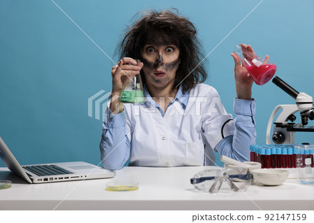 Mad silly looking crazy lab worker sitting at desk while having glass jars filled with experimental liquid compounds. Foolish scientist with funny face expression handling beakers filled with serum. Mad silly looking crazy lab worker sitting at desk while having glass jars filled with experimental liquid compounds. Foolish scientist with funny face expression handling beakers filled with serum. 92147159